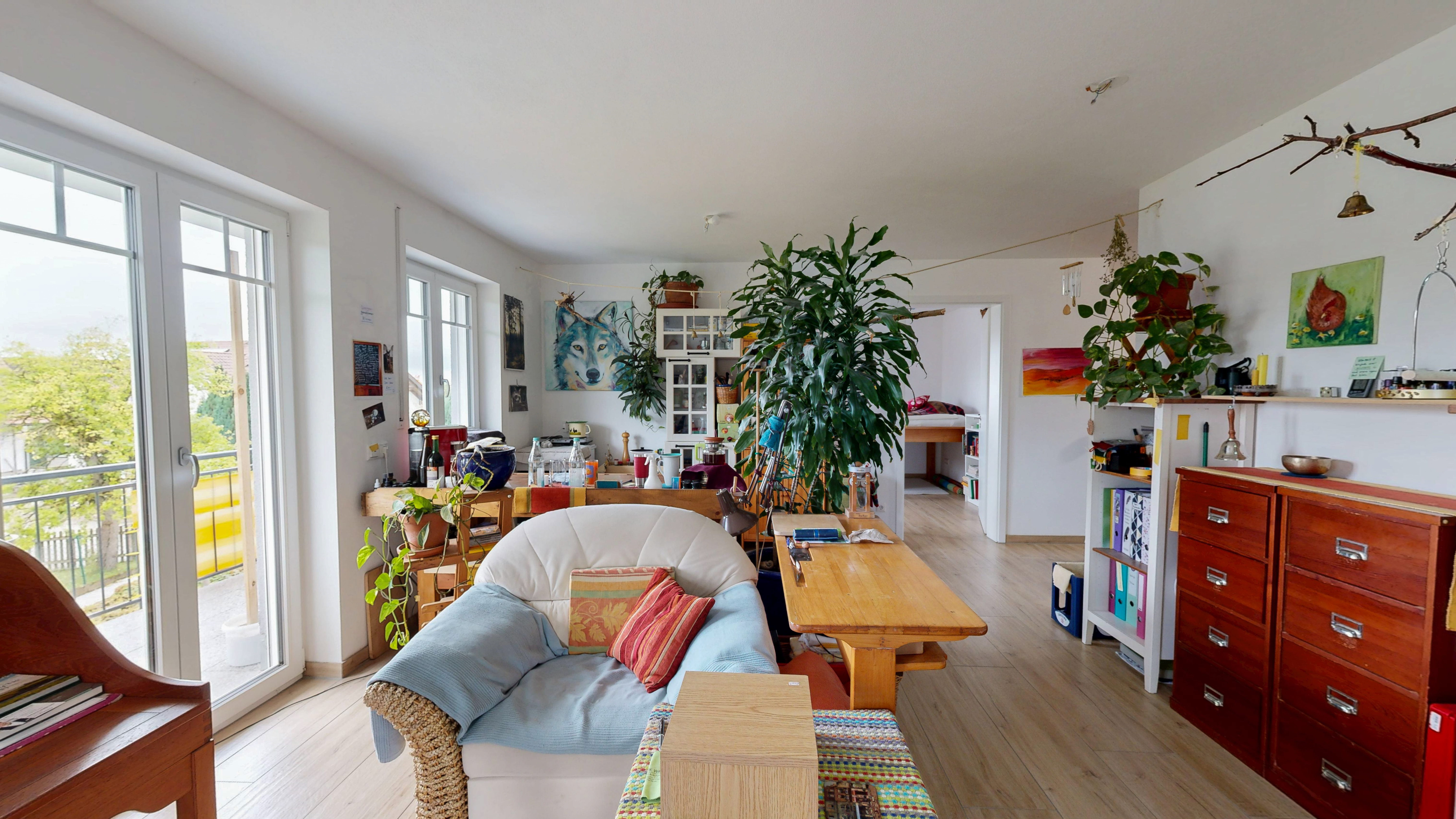 Vorher-Bild für virtuelles Home Staging und digitales Entrümpeln (Decluttering). Ein unruhiger, stark zugestellter Wohnbereich mit sehr persönlicher Einrichtung, großen Zimmerpflanzen und dominierenden Holzmöbeln. Ein Sessel im Vordergrund, Tische und Regale voller Alltagsgegenstände lassen den Raum optisch kleiner wirken. Große Balkontüren auf der linken Seite lassen zwar viel Tageslicht herein, jedoch lenkt die Unordnung vom eigentlichen Schnitt ab. Ein klassisches Beispiel für ein Objektbild, dessen wahres Potenzial und Raumgröße erst durch eine professionelle digitale Neu-Möblierung für die Immobilienvermarktung sichtbar gemacht werden muss.